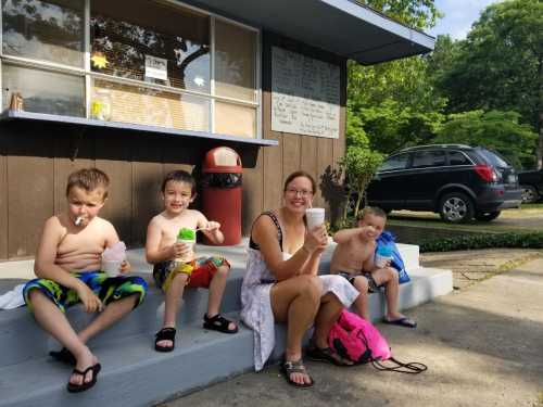 A woman and three boys sit on steps, enjoying colorful drinks on a sunny day outside a small building.