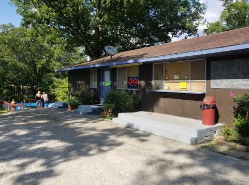 A small brown building with an "OPEN" sign, surrounded by trees and a gravel path, with people in the background.