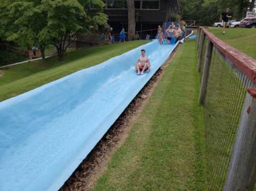 A water slide with children sliding down on a sunny day, surrounded by green grass and trees.