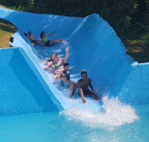 A group of people joyfully sliding down a blue water slide, splashing into the pool below on a sunny day.