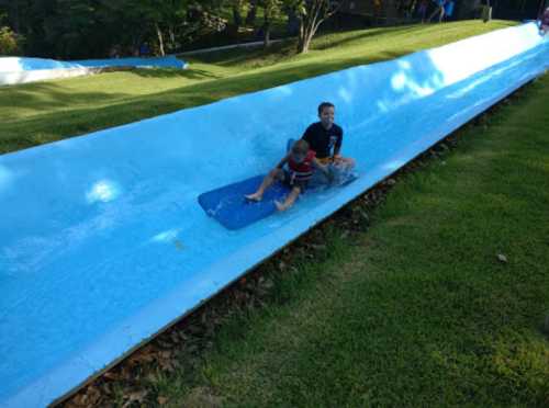 Two children slide down a blue water slide, surrounded by green grass and trees.