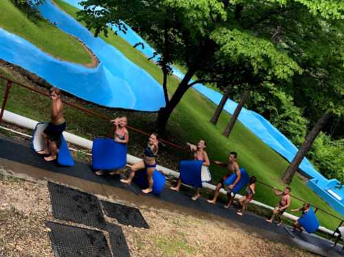 A group of people carrying blue inflatable mats walking up a path beside a water slide in a lush, green park.