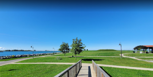 A sunny park with green grass, trees, and a pathway leading to a waterfront with boats in the distance.