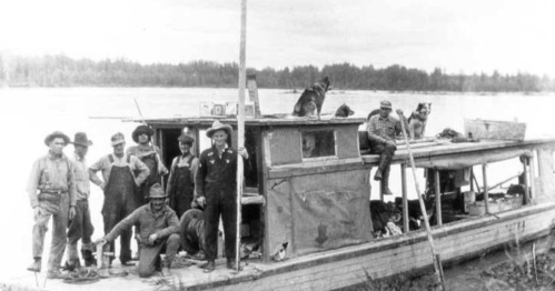 A group of men and a dog pose on a vintage boat by a river, surrounded by trees in the background.
