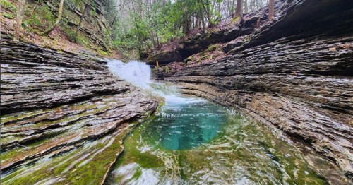 A serene natural scene featuring a small waterfall cascading into a clear pool surrounded by rocky cliffs and lush greenery.