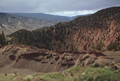 A rugged landscape featuring layered rock formations and green vegetation against a backdrop of mountains and cloudy skies.