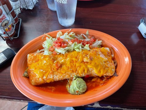 A large enchilada topped with melted cheese, served with lettuce, tomatoes, and guacamole on a vibrant orange plate.