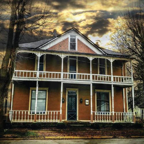 A vintage two-story house with a porch, set against a dramatic cloudy sky. Trees frame the scene.