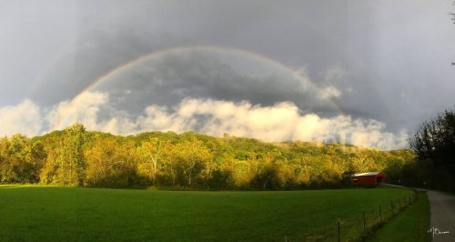 A vibrant rainbow arcs over a lush green field and wooded hills, with a red barn visible in the distance.