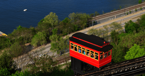 A red and black funicular car travels along tracks above a river, surrounded by trees and a road below.