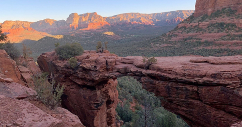 A stunning rock arch in a desert landscape, surrounded by red cliffs and green trees under a clear blue sky.