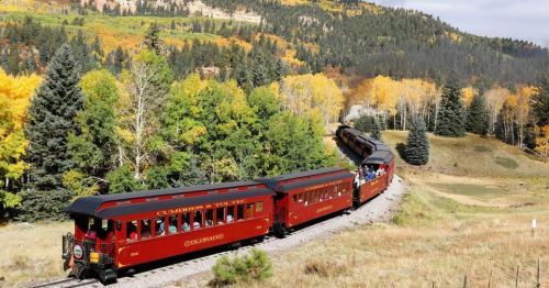A red train travels along a scenic route surrounded by colorful autumn trees and rolling hills.