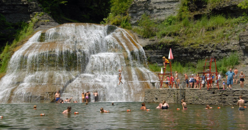 A waterfall cascades into a swimming area with people swimming and lounging on the rocks in a sunny setting.