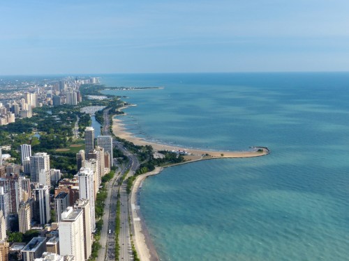 Aerial view of a coastal city with sandy beaches and a blue lake, surrounded by tall buildings and greenery.