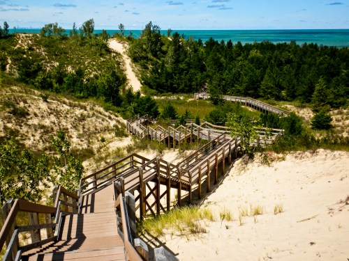 A wooden boardwalk winds through sandy dunes and greenery, leading to a serene blue lake in the background.