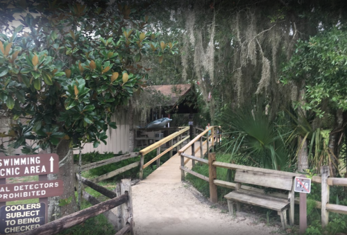 A wooden path leads to a picnic area surrounded by trees and greenery, with signs indicating rules and directions.