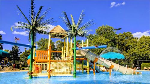 Colorful water play area with slides, palm trees, and a thatched roof, set against a bright blue sky.