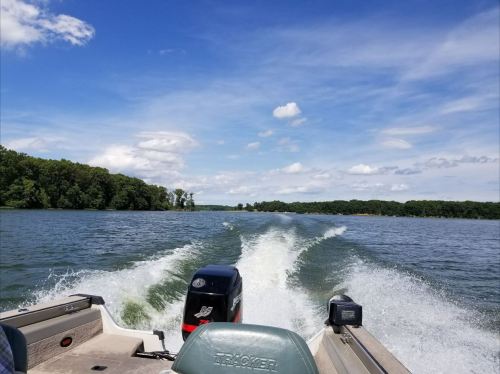 A boat's wake trails behind as it moves across a calm lake under a blue sky with scattered clouds.