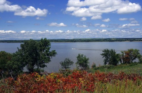 A scenic view of a river with lush greenery and colorful foliage under a blue sky with fluffy clouds.