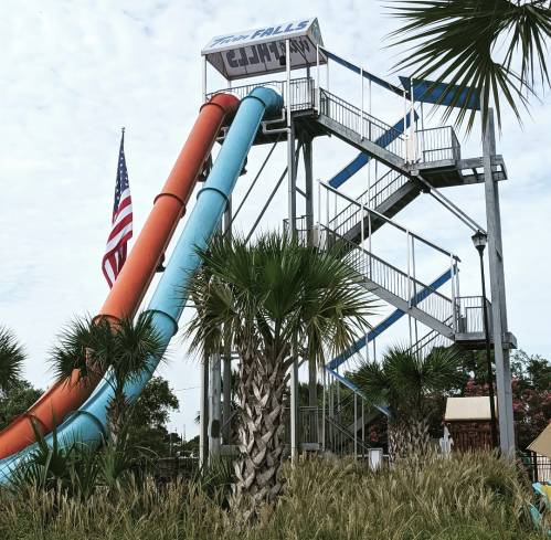 A tall water slide structure with orange and blue slides, surrounded by palm trees and an American flag.