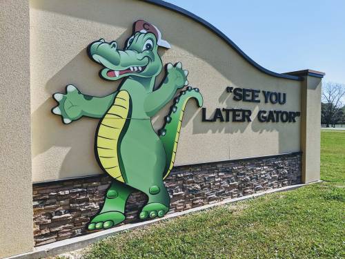 A cartoon alligator wearing a cap, next to the phrase "See You Later Gator" on a sign. Green grass in the foreground.