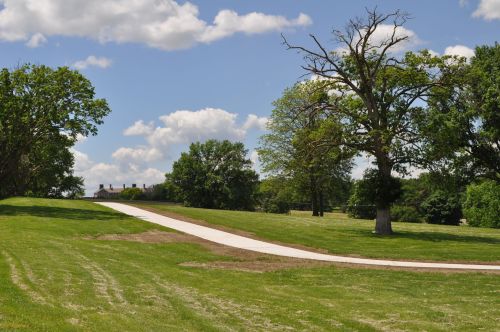 A winding path through a grassy landscape with trees under a partly cloudy sky. A building is visible in the distance.