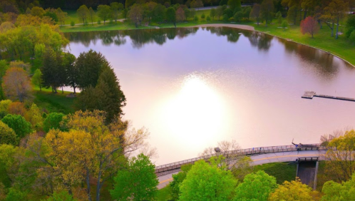 A serene lake surrounded by lush greenery, reflecting sunlight, with a winding path and trees in the background.