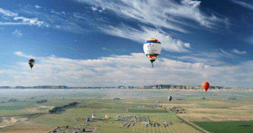 Colorful hot air balloons float over a vast green landscape under a blue sky with wispy clouds.