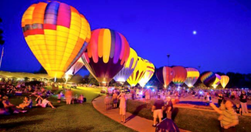 Colorful hot air balloons illuminated at night, with a crowd enjoying the event on a grassy field.