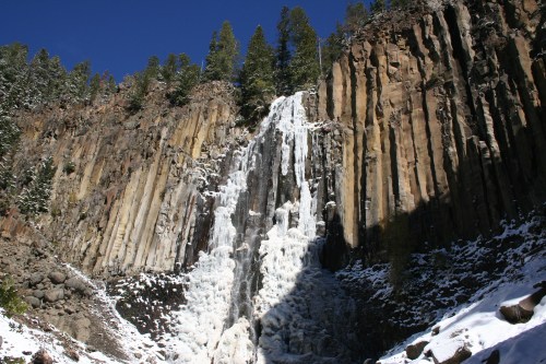 A frozen waterfall cascading down rocky cliffs, surrounded by snow-covered ground and evergreen trees under a clear blue sky.