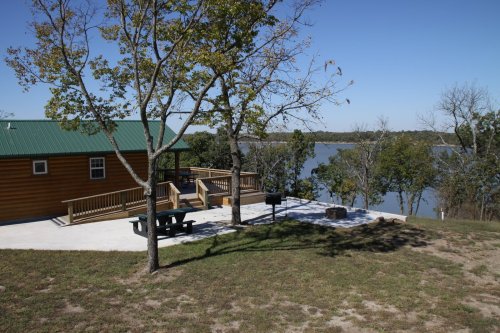 A wooden cabin by a lake, surrounded by trees, with a picnic area and grill on a sunny day.