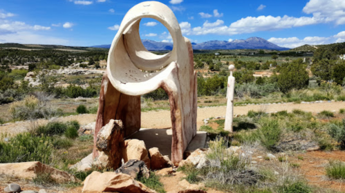 A large, abstract stone sculpture with a circular opening, set against a backdrop of mountains and blue sky.