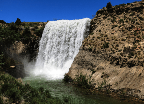 A cascading waterfall flows over a rocky cliff into a serene green pool, surrounded by lush vegetation and clear blue skies.