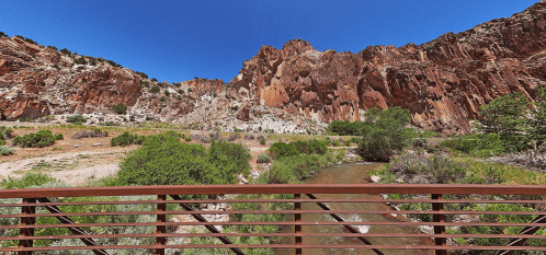 A scenic view of rocky cliffs and a flowing stream, framed by lush greenery under a clear blue sky.