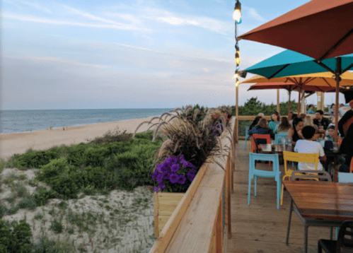 A beachside restaurant with colorful umbrellas, diners enjoying the view, and a sandy shore in the background.