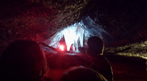 A dimly lit cave with stalactites illuminated by red and white lights, with silhouettes of people in the foreground.
