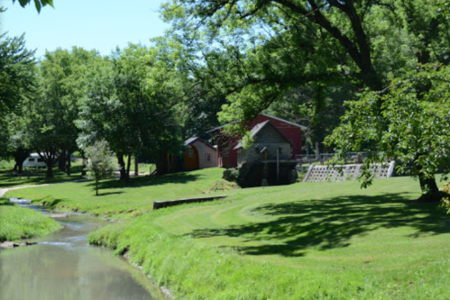 A serene landscape featuring a creek, lush green grass, and a rustic red barn surrounded by trees.