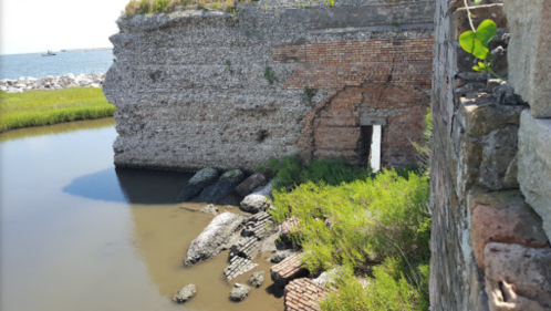 Ruins of a brick structure by a calm waterway, surrounded by green grass and rocky outcrops.