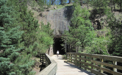 A person walks on a wooden bridge leading to a dark tunnel surrounded by trees and rocky cliffs.