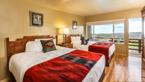 Cozy hotel room with two beds, colorful blankets, wooden furniture, and large windows showcasing a scenic view.