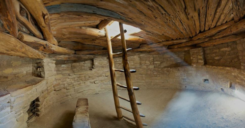 Interior of a rustic, circular room with wooden beams, a ladder, and a skylight, featuring stone walls and a sandy floor.
