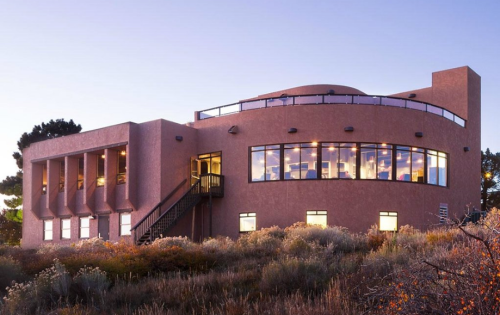 A modern, rounded building with large windows, surrounded by shrubs and grass, at dusk.