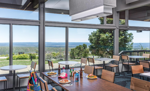 Bright dining area with large windows showcasing a scenic view, featuring tables set with plates of food and drinks.