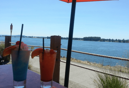 Two colorful cocktails sit on a table with a scenic view of a river and trees under a clear blue sky.