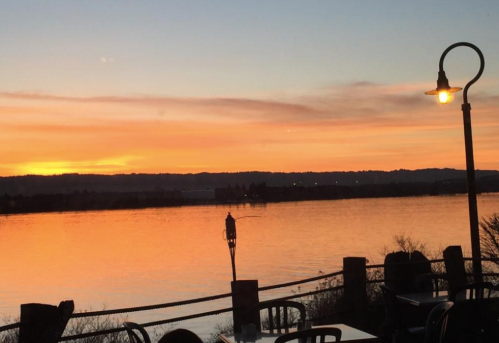 A serene sunset over a calm river, with warm orange and pink hues reflecting on the water and a lamp post in the foreground.