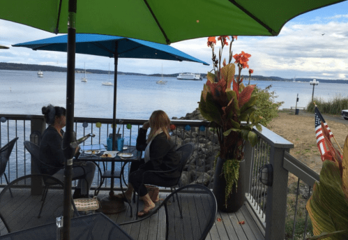 Two people sit at a table under umbrellas, enjoying a meal with a view of the water and boats in the distance.