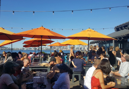 A lively outdoor dining area with orange umbrellas, filled with people enjoying food and drinks against a sunny backdrop.