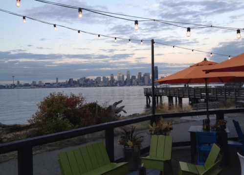 View of a waterfront with city skyline, string lights, and colorful patio furniture at dusk.