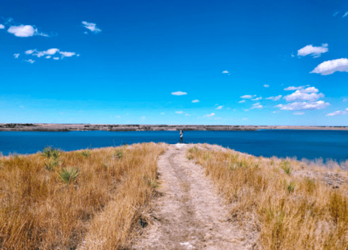 A person stands on a grassy path by a calm blue lake under a clear blue sky with scattered clouds.