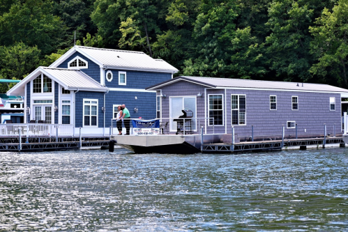 Two colorful houses on stilts by the water, with a small boat docked nearby and lush greenery in the background.